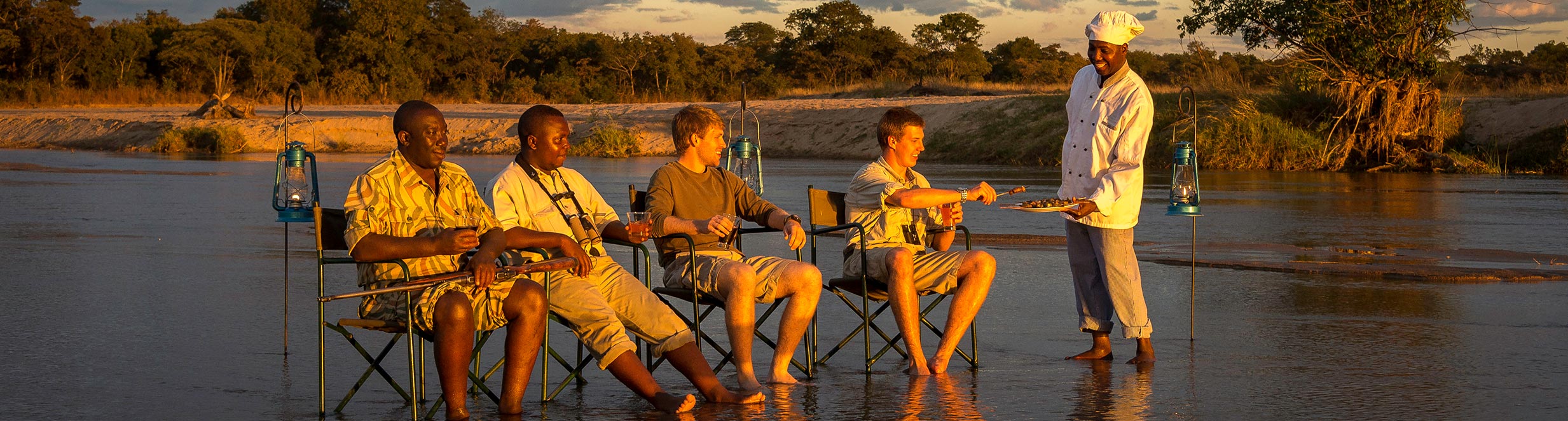 Several people sitting in chairs in a river being served snacks by a waiter
