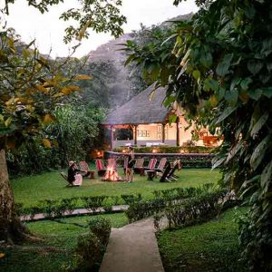 View of the lodge from the forest with campfire on a lawn surrounded by chairs